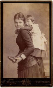 Sepia photograph, an 1880s woman wearing a dark full-length dress gives a piggy back ride to a girl wearing boots and a white apron over her dark dress.